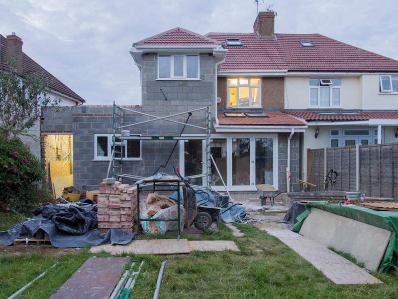 A semi-detached house with a two-storey side and rear extension in blockwork, a scaffold tower and materials on the lawn