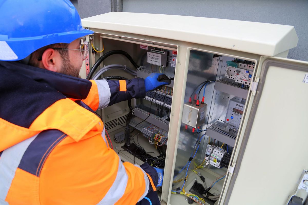 An electrician in a blue hard hat, safety glasses, gloves and a high-visibility jacket works on the wiring inside an open distribution cabinet